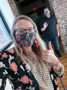 Image of Jolinda in mask with man (Jonathan) in background waving.