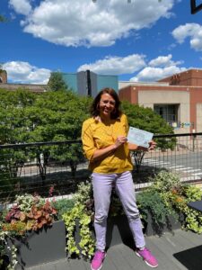 Jolinda in a yellow shirt and pink shoes on a roof with a certificate.