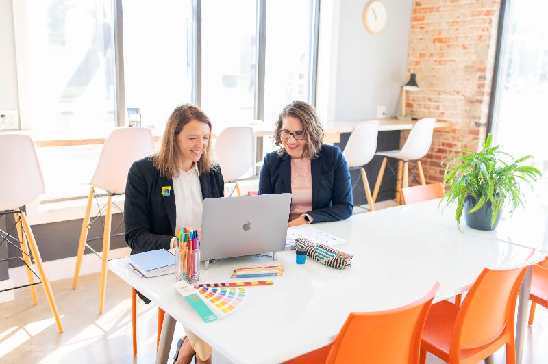 Image of two women sitting at a table and looking at a computer while in an office space.